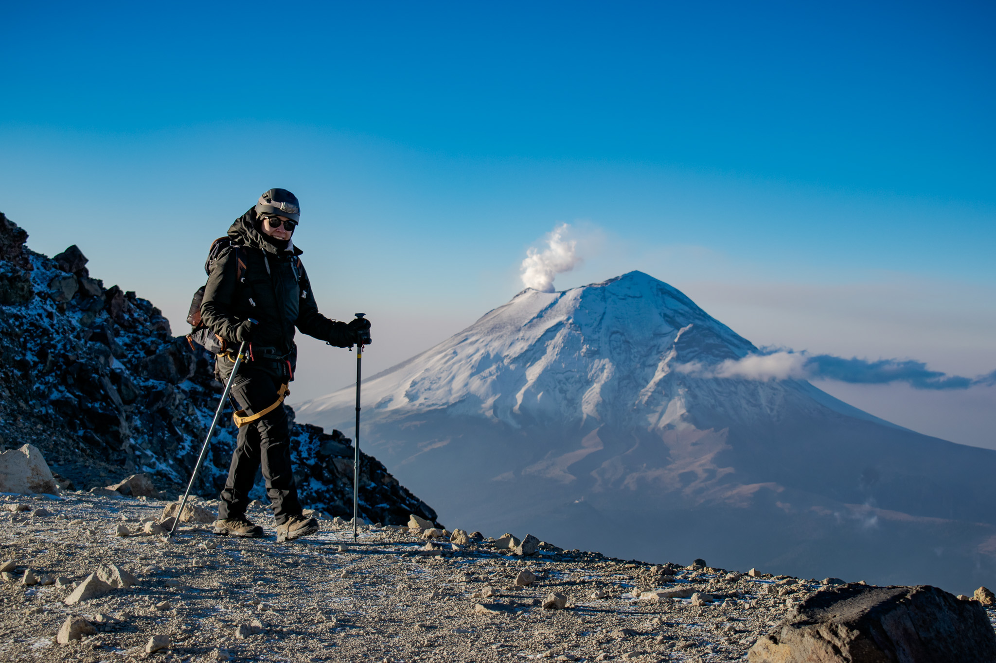 Climber on Pico de Orizaba summit with Popocatépetl erupting steam under a deep blue sky