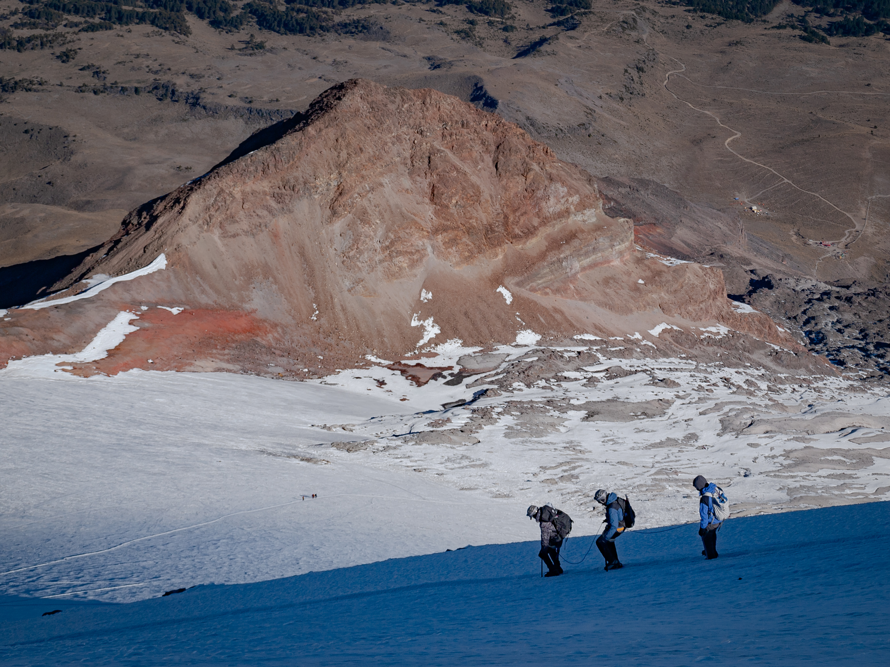 Three climbers on the glacier with a rust-red volcanic formation rising behind