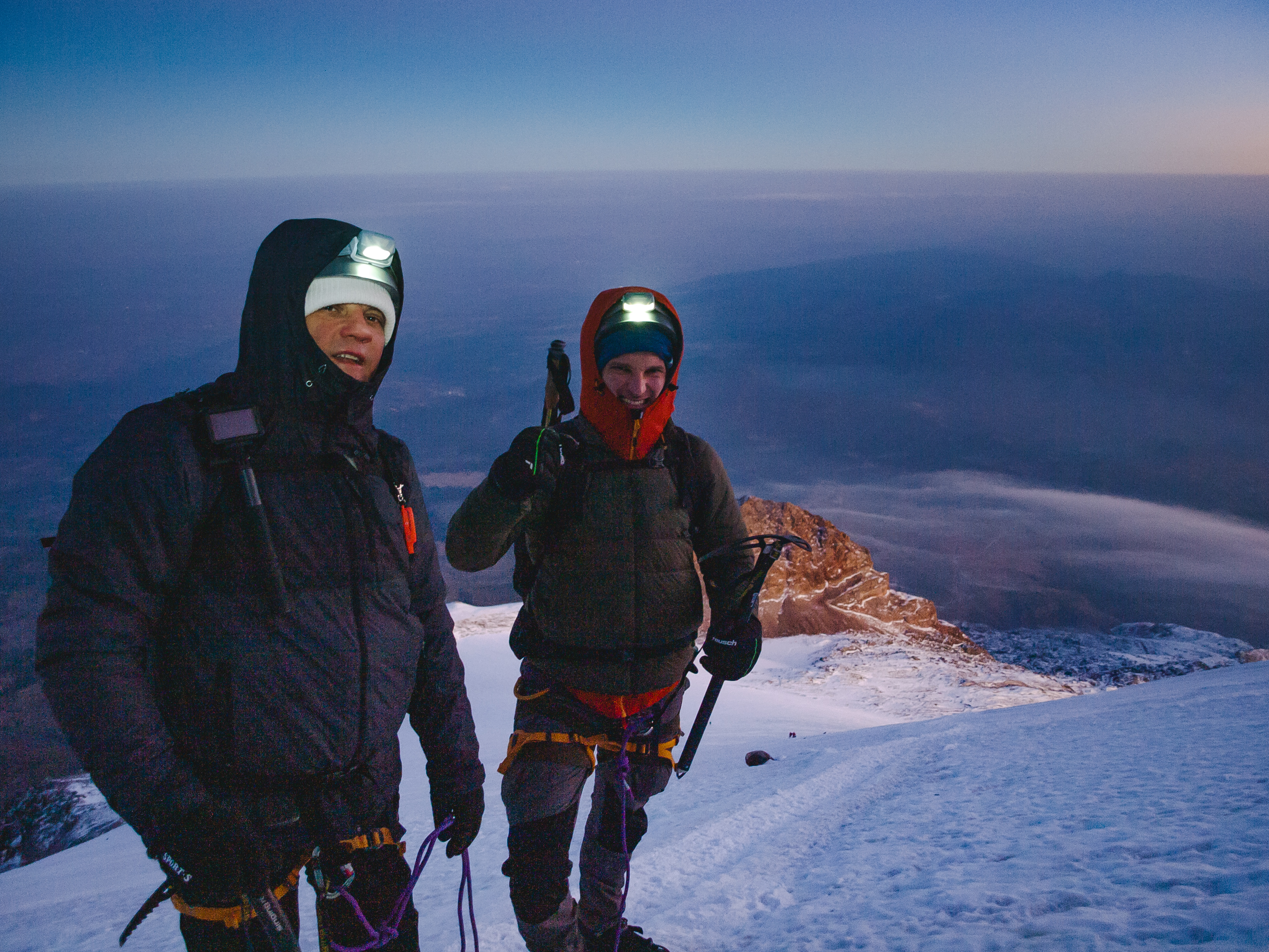 Two climbers at the Pico de Orizaba summit with headlamps still on — dawn breaking pink and orange behind them