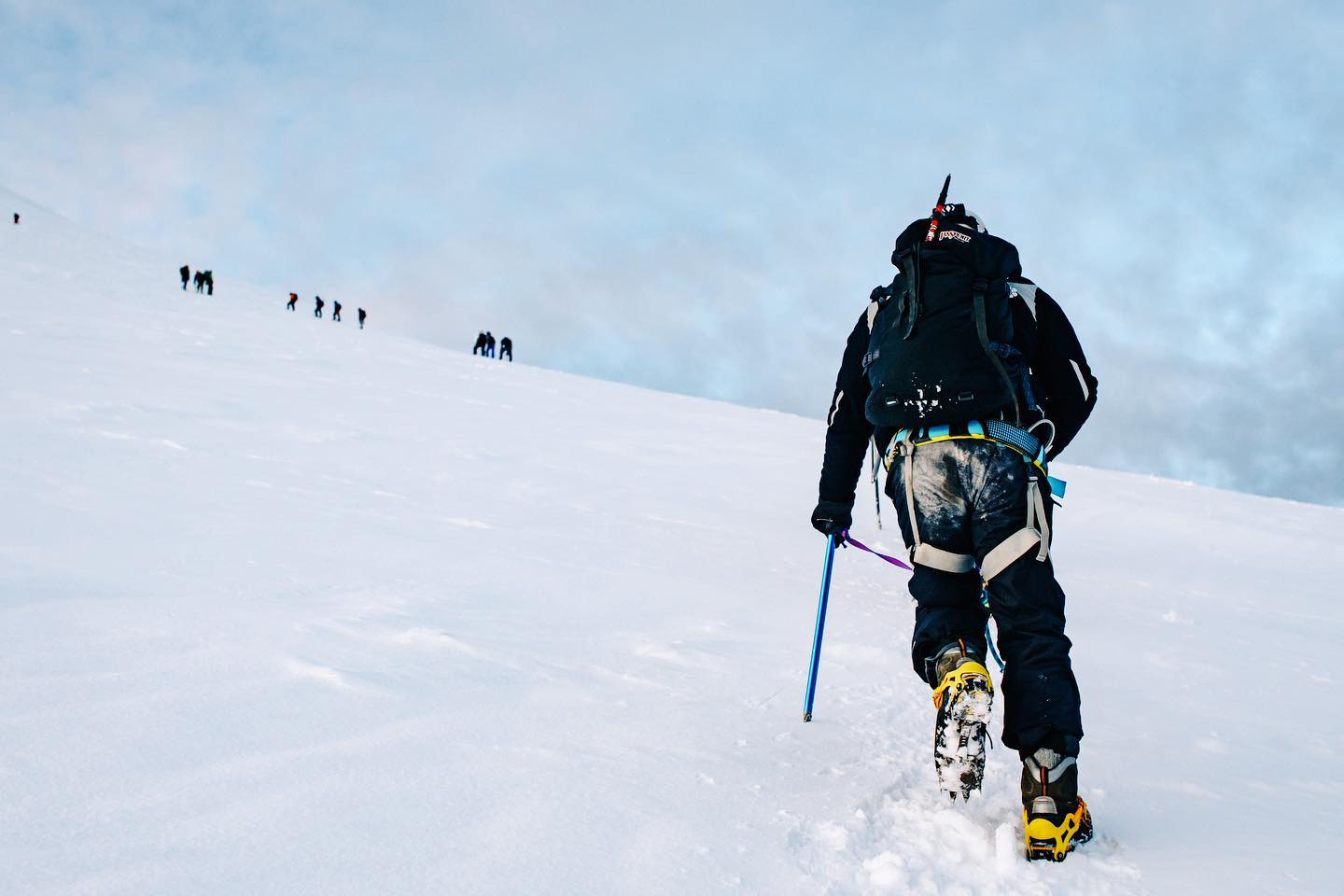 Climber in crampons and harness ascending the steep glacier — teammates visible behind