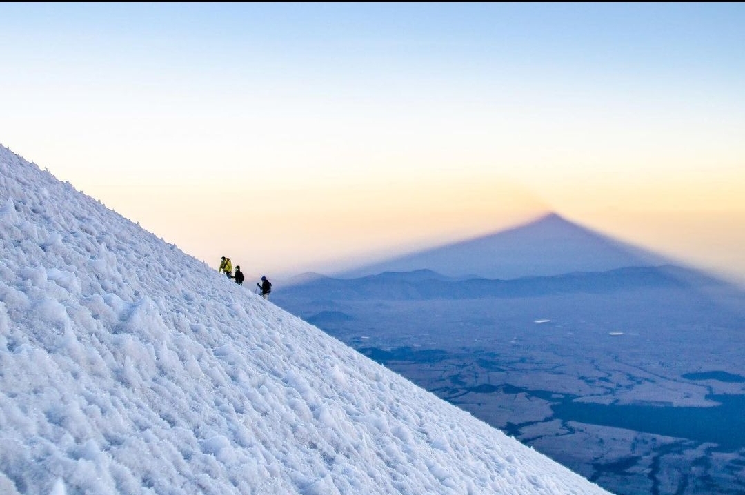 Two climbers on the Pico de Orizaba icefall at first light — the mountain's own shadow stretches across Mexico below