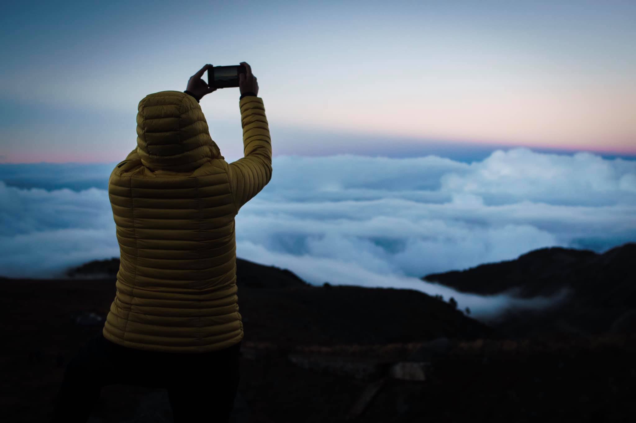 Climber silhouetted against a sea of clouds at dusk, phone raised to shoot the view